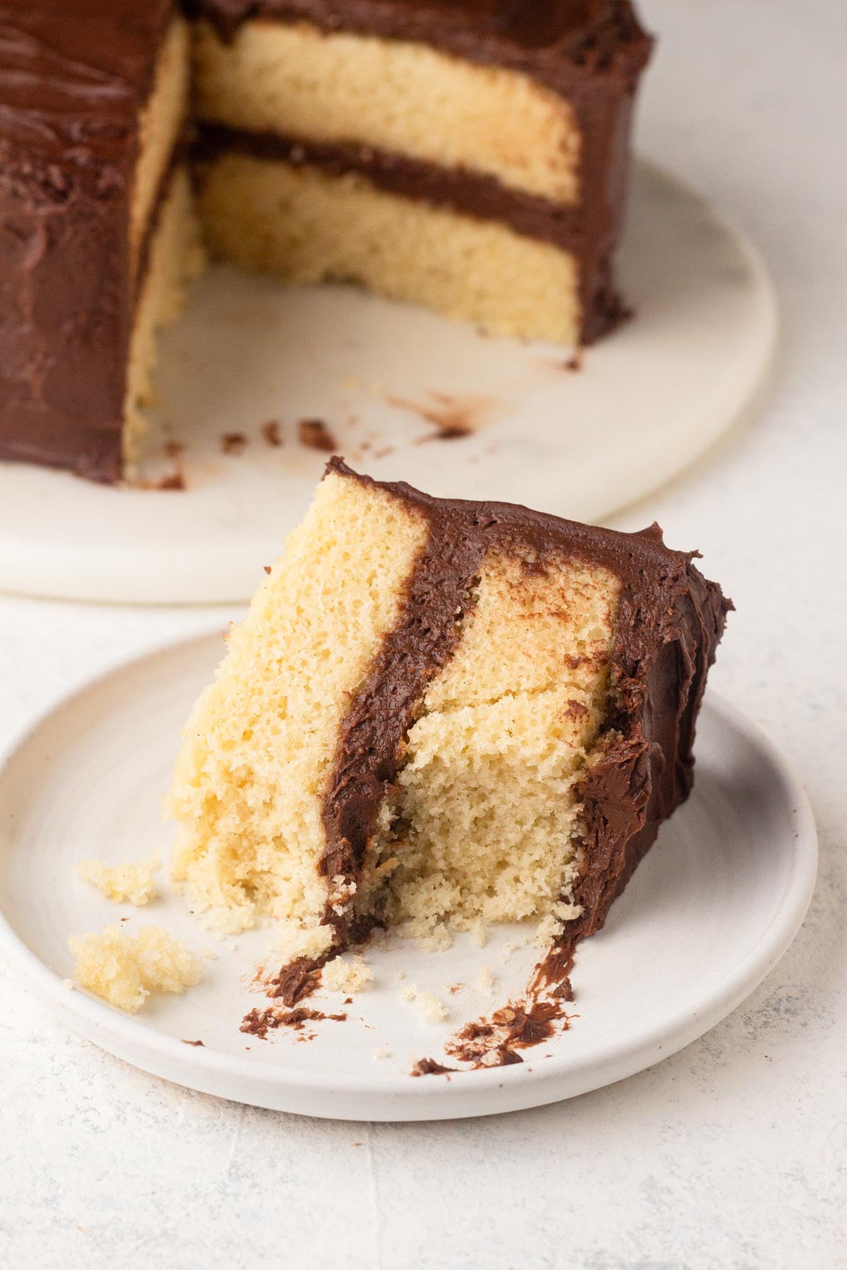 A partially-eaten slice of vanilla layer cake with chocolate frosting on a white plate.