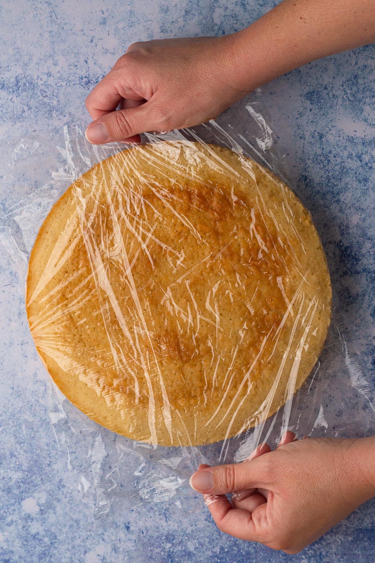 Hands wrapping a round vanilla cake layer with plastic wrap.