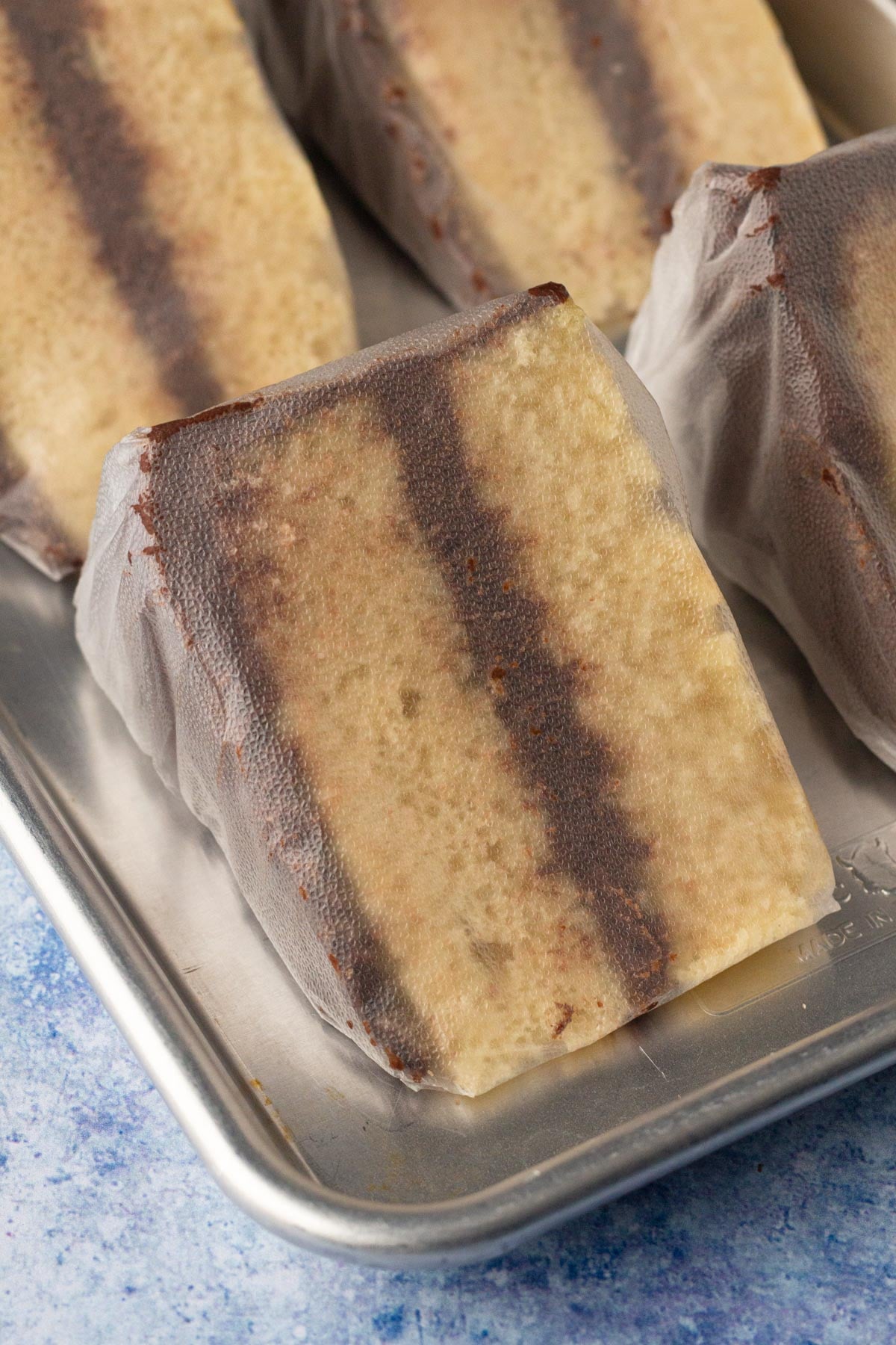 Slices of vanilla cake with chocolate frosting wrapped in plastic wrap on a baking sheet.