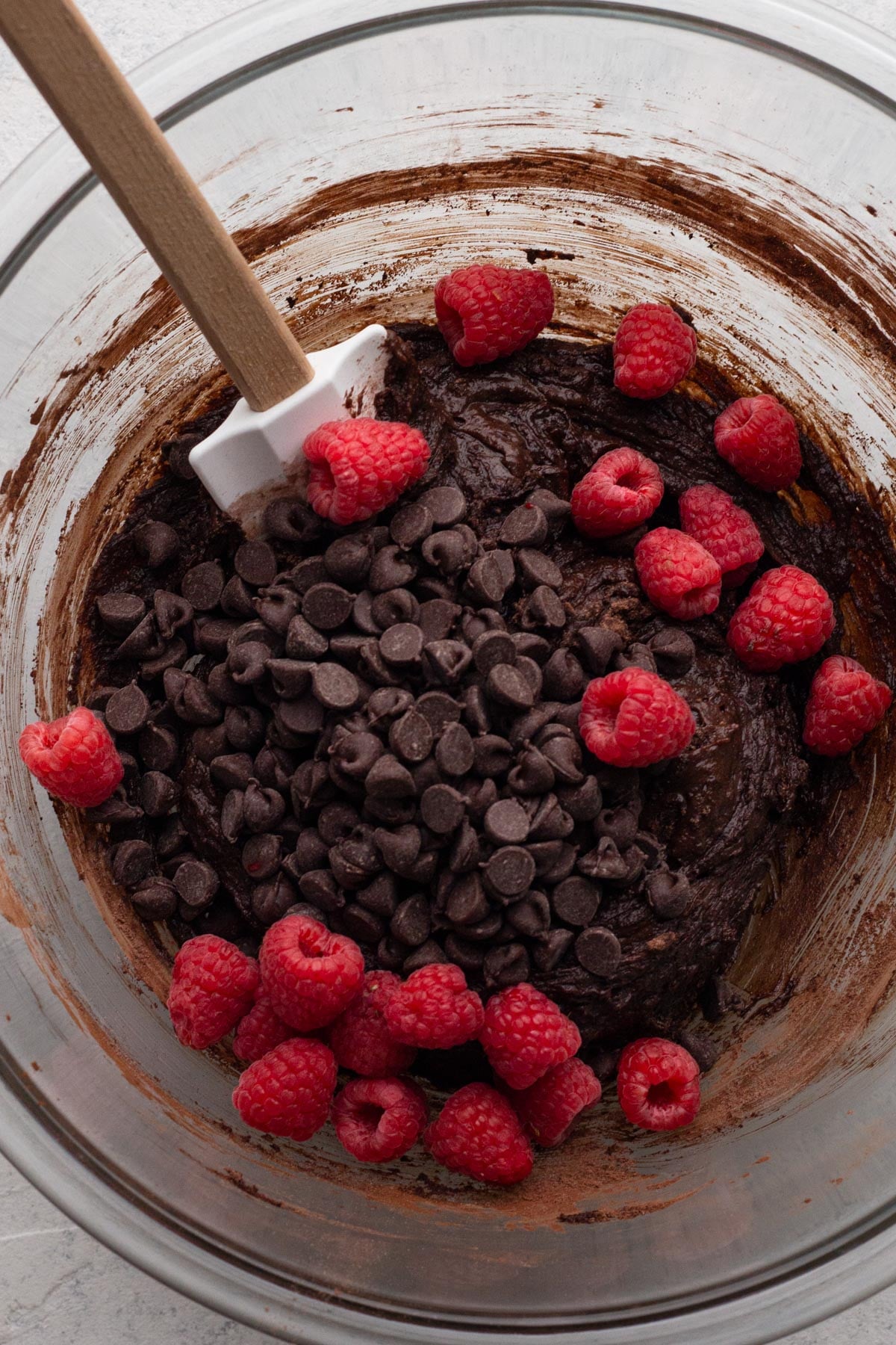 Brownie batter with fresh raspberries and chocolate chips in a glass mixing bowl with a rubber spatula.