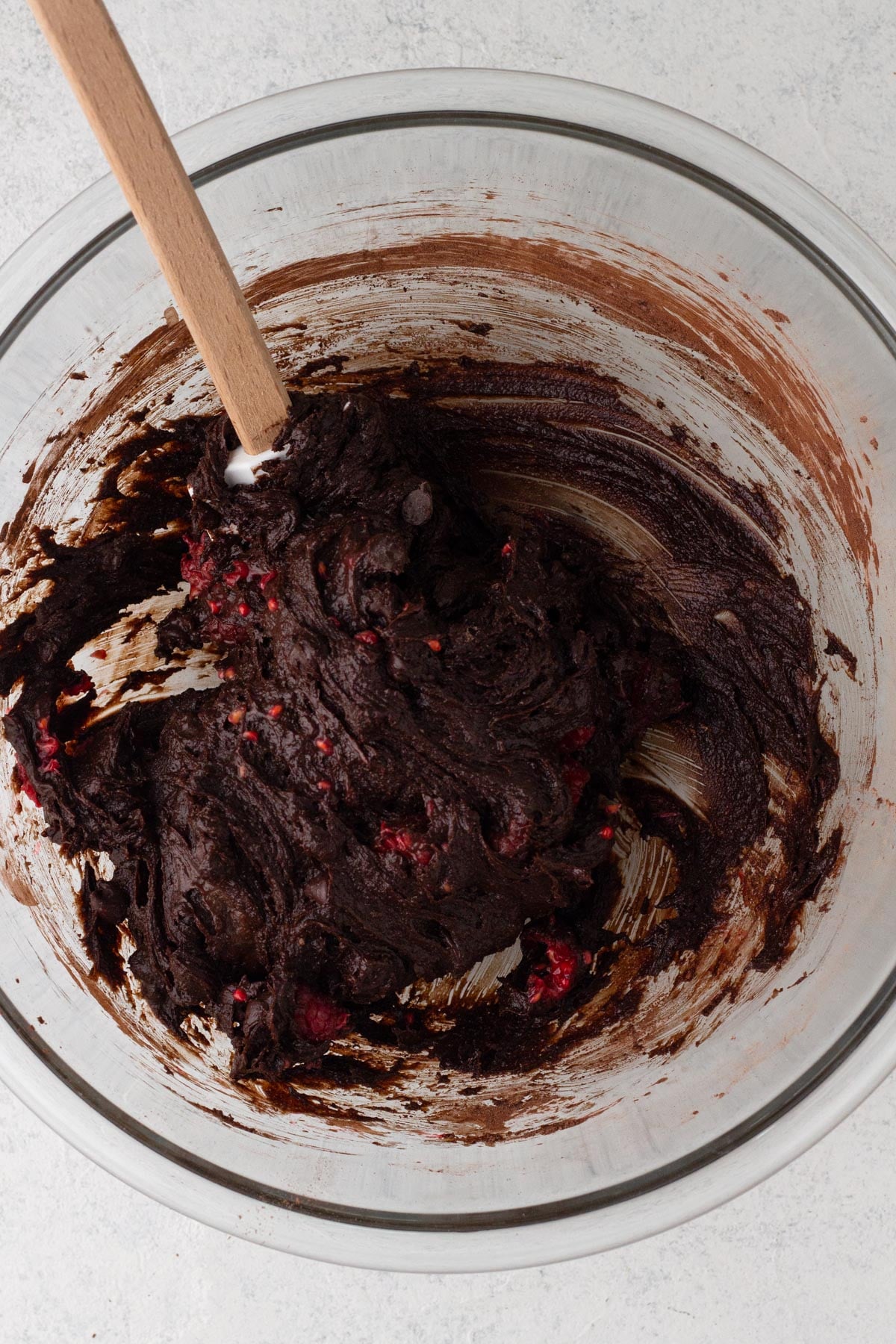 Brownie batter with raspberries in a glass mixing bowl with a rubber spatula.